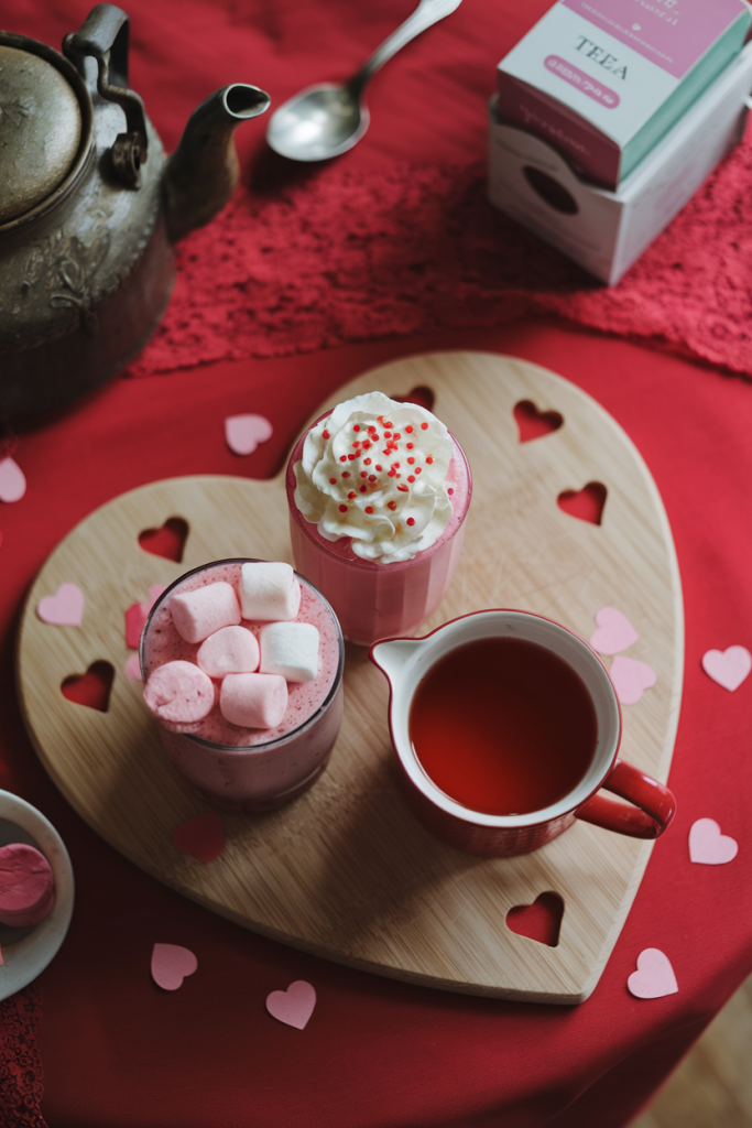 A cozy trio of hot Valentine's Day drinks featuring steaming mugs of strawberry hot chocolate, chocolate cherry bliss, and hibiscus honey tea, with whipped cream and chocolate garnishes