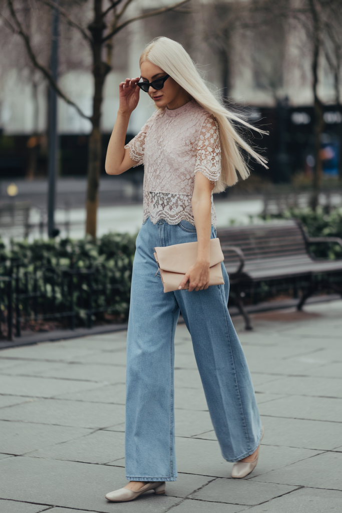 woman wearing a pastel lacey top with jeans and ballet flats waling in a sunny park.